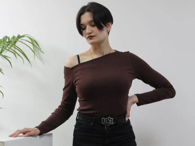 KalipsoTryi with short black hair, brown off-shoulder top, black belt with buckle, black pants, standing with arm on white surface, plant in background, small earrings, confident pose.