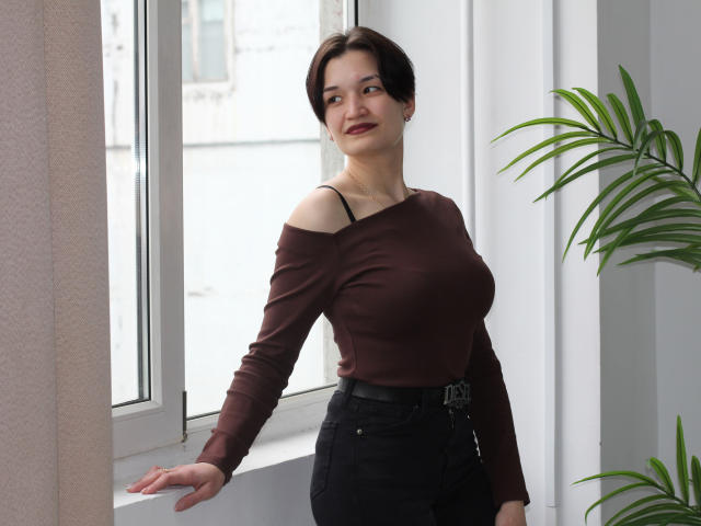 KalipsoTryi with short brown hair, wearing a brown off-shoulder top, black high-waisted pants, standing by a window, green potted plant on the right, smiling, hands resting.