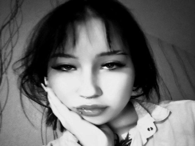Black-and-white photo of RosemarySchwall with dark, messy hair, heavy eyeliner, nose piercing, lip piercing, and hand on cheek, wearing a button-up shirt, looking slightly down.