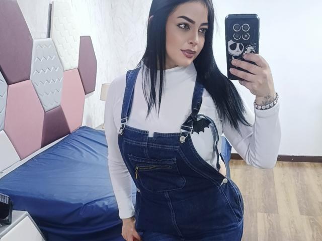 LilithAmoth, avec de longs cheveux noirs, portant une salopette en jean, un col roulé blanc et un t-shirt avec des chauves-souris, prend un selfie devant un miroir, montrant ses seins et ses fesses, dans une pièce avec un lit..