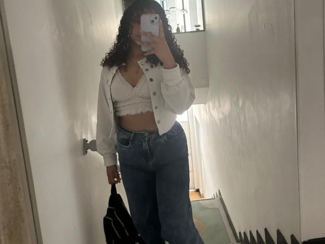 Sofia, mit lockigen Haaren, weißem Spitzen-BH, Hemd und hoch taillierten blauen Jeans, hält ein Handy im Spiegel-Selfie, eine schwarze Tasche liegt auf dem Boden, im Hintergrund weiße Wände..