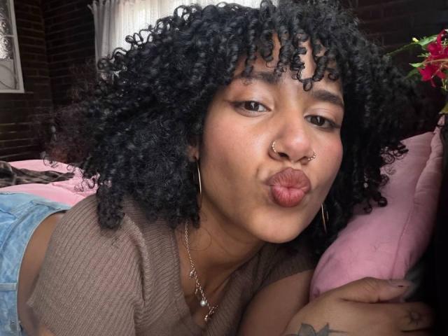 CinthiaWalker with curly black hair, brown skin, nose ring, and hoop earrings, puckering her lips in a kissy face. She's wearing a brown top and blue denim shorts.