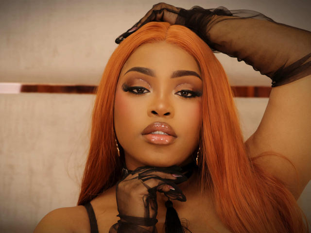 TiannahGlow with orange hair, glossy lips, black sheer gloves, and gold earrings, raising her right arm, showing her tits and pussy hint, in a sexy, confident pose.