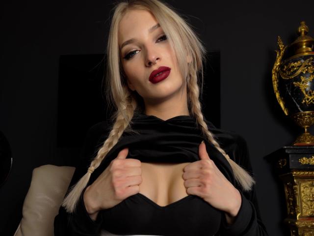 FairyFox-hot with blonde braids, red lipstick, and black velvet top, pulling down to show her big tits, in a dark room with ornate golden decor on the right.