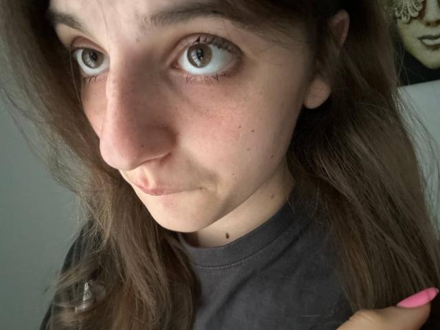 KarieMestler's face with wide eyes, brown wavy hair, pink lips, small nose, and freckles. She's wearing a dark shirt, with a pink nail visible on her right hand.