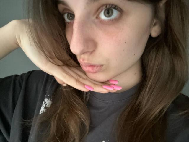 KarieMestler with pink nails, pouty lips, and brown hair, wearing a black shirt, touching her cheek with her hand, grey eyes, and a few freckles on her face.