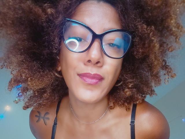 CinnamonBabe, avec ses cheveux bruns bouclés, ses lunettes noires, son rouge à lèvres rose et son haut noir à bretelles, montre un tatouage de dragon sur son épaule droite, et sourit à l'appareil photo..