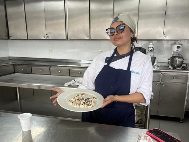 GalaMalkova, con gli occhiali e un foulard bianco in testa, tiene un piatto con pezzi di funghi in una cucina, indossa un grembiule blu sopra una camicia bianca, e sorride..