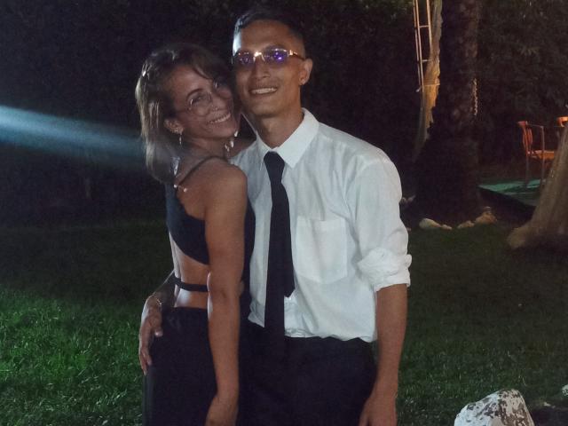 AlejoyMaria with tits in a black crop top and pants, standing next to a dude in a white shirt and black tie, both smiling, in a grassy night setting.