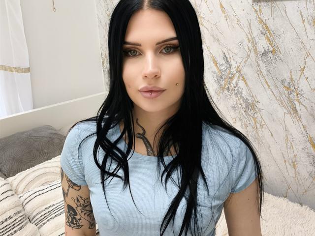 TattooDolly con cabello negro largo, ojos verdes, camisa azul claro, tatuajes en los brazos y una pequeña marca de nacimiento en la mejilla, sentada en una cama..