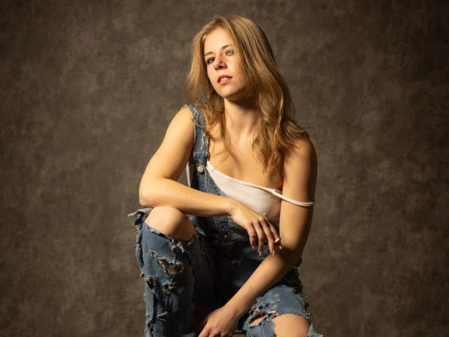 Isadora-hot with long blonde hair, wearing torn blue jeans and a white top slipping off her shoulder, sits against a dark textured background, looking slightly up and to the side.
