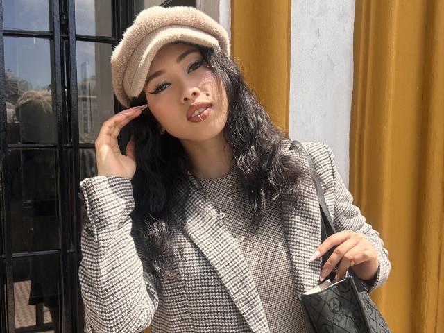 NashiroSakuraii with long black hair, beige beret, checkered shirt, and yellow curtains, licking her glossy lips, holding a black bag, touching her face, looking sexy.