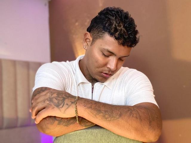 AxellKing69 con i capelli ricci, camicia bianca, braccia tatuate, braccia incrociate, che guarda in basso, braccialetto dorato, sedia verde, sfondo marrone..