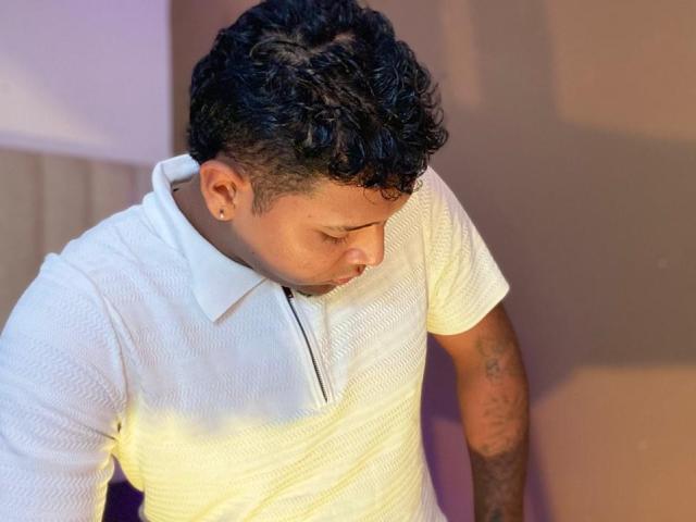 AxellKing69, avec ses cheveux noirs bouclés, porte une chemise polo blanche texturée, regarde vers le bas, a des tatouages sur son bras droit et un petit anneau de nez sur sa narine gauche..