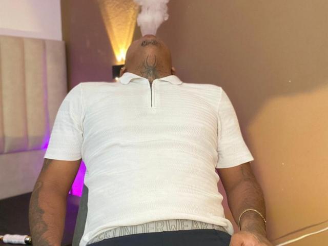 AxellKing69 es un tipo calvo, con tatuajes, que lleva una camisa polo blanca, está fumando un porro y está recostado con el trasero levantado, los pantalones ligeramente bajados, revelando su cinturón..
