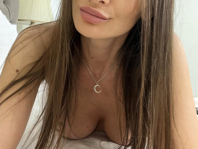 AliceMarr with long brown hair, pink lips, and a silver necklace, leaning forward, showing her big tits and cleavage, on a white bed.