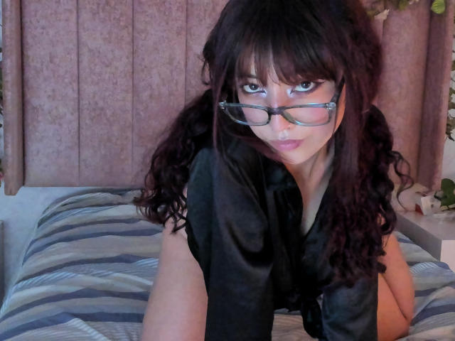 LilyDickenson with curly black hair, wearing glasses, and a black silky shirt, leaning on a bed with striped sheets, looking seductively at the camera.