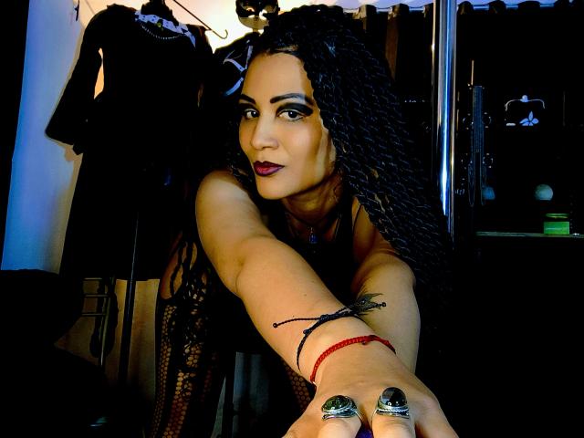 DivaMarie mit langen, schwarzen Zöpfen, dunklem Lippenstift, die nach vorne blickt, mit einem roten Armband, einem schwarzen Tattoo am Arm, einem grünen Ring und dunklen Nägeln, die in einen schwach beleuchteten Raum schaut..