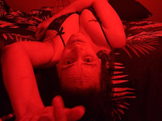 MiaWowx ligt op een bed, gekleed in zwarte kant lingerie, met rood licht, waarbij haar borsten zichtbaar zijn, haar armen zijn omhoog, ze kijkt omhoog en maakt een "kom hier" gebaar met haar vingers..
