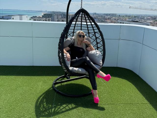 MoniqueBliss with blonde hair, sunglasses, black top, tight black pants, and pink heels, sitting in a black egg chair on green turf, city skyline in the background.