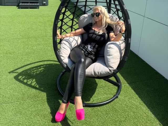 MoniqueBliss with big boobs, wearing black leather pants, black top, pink heels, holding a phone, sitting in a black hanging chair on green grass, wearing black sunglasses.