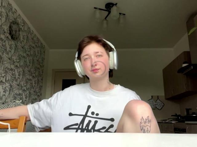 KristelHenandez, a pale-skinned woman with short brown hair, wearing white headphones and a white t-shirt, sits with a tattoo on her knee, pouting slightly.