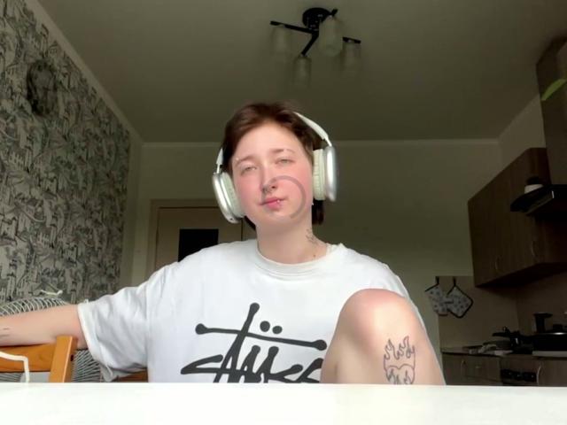 Kristel Henandez, avec des cheveux bruns courts, des écouteurs blancs, une chemise blanche, des lèvres pulpeuses, un tatouage sur le genou droit, assise, regardant la caméra, avec un fond de cuisine et de la lumière provenant d'une fenêtre..