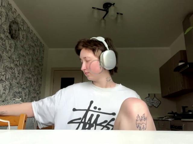 KristelHenandez with white headphones, pink cheek, white shirt, and tattoo on knee, sitting with knee up, short brown hair, in a room with a patterned wall.