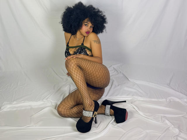 LaCoquineX, avec ses cheveux noirs bouclés, son soutien-gorge en dentelle vert foncé, ses bas résille, ses escarpins noirs à semelles rouges, assise sur un tissu blanc, montrant ses fesses et son bras tatoué..