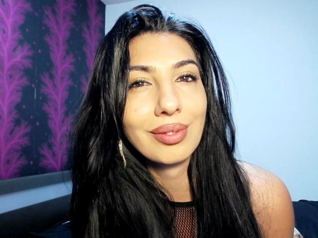 A BrunetteBabe69 tem cabelos pretos longos, olhos castanhos e lábios rosados cheios com um leve sorriso. Ela está vestindo uma blusa de malha preta e tem brincos dourados..