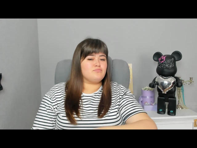 CharlotaTilb with brown hair and bangs, wearing a striped shirt, sitting with arms crossed, next to a black Minnie Mouse doll with a heart and pink bow.