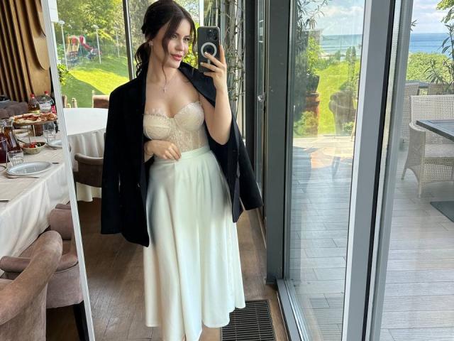 RaichelStoun with tits in a white lace strapless top, black jacket, and white skirt, taking a mirror selfie. She's standing by a window, showing a beach view outside.