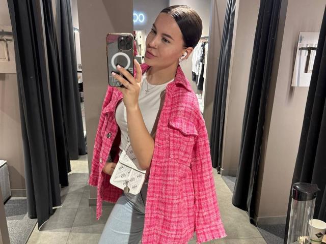 RaichelStoun in a pink plaid jacket, white t-shirt, and gray jeans, holding a phone in a mirror selfie, with brown hair slicked back, and a confident smile.