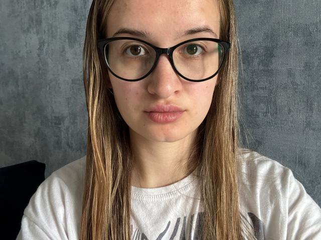 Beverlee Belyea, met rechtbruin haar, zwarte brillen en een wit T-shirt, heeft een neutrale uitdrukking en licht gezwollen lippen. Haar groene ogen zijn gecentreerd in het beeld..