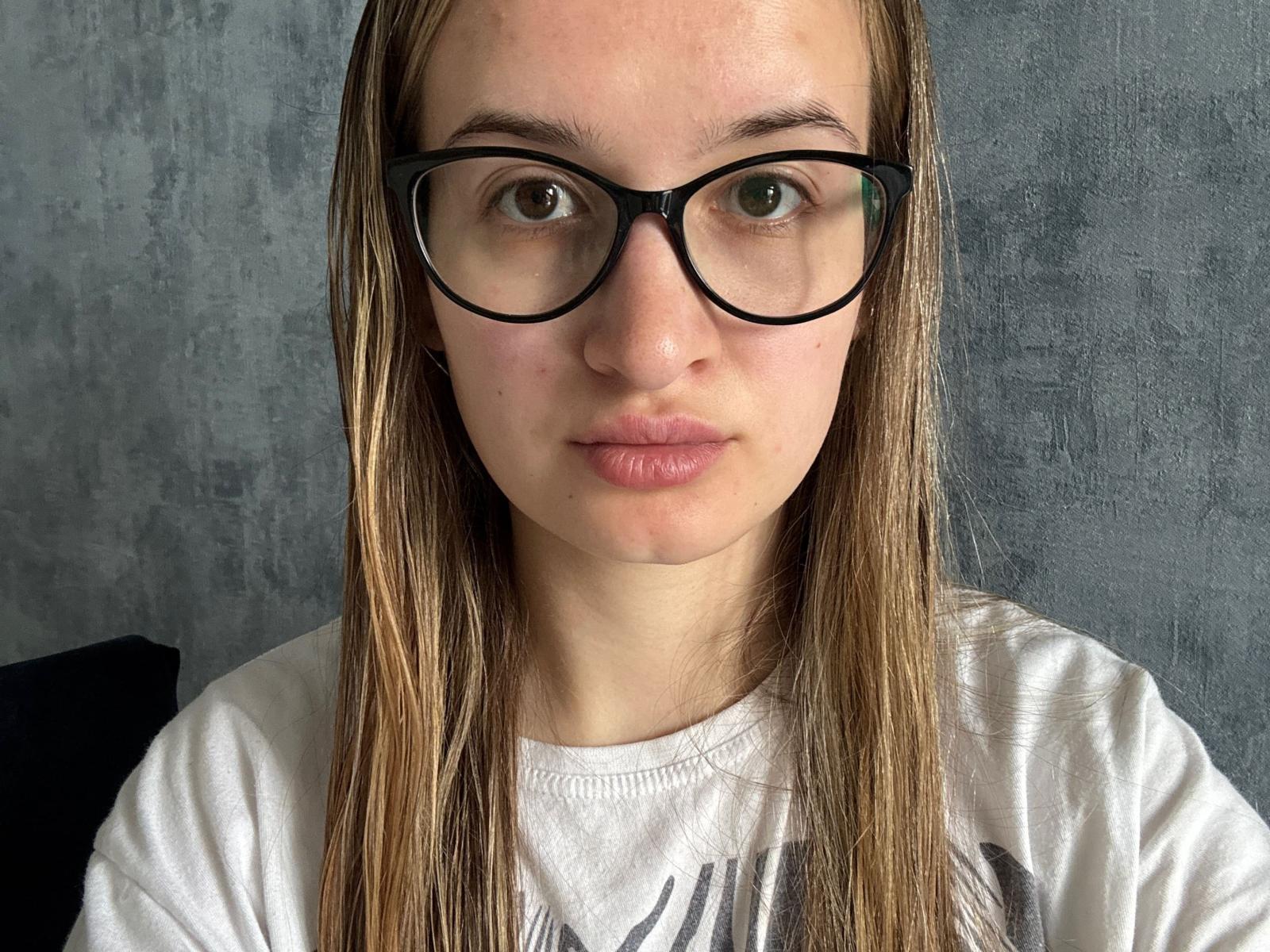 Beverlee Belyea, met rechtbruin haar, zwarte brillen en een wit T-shirt, heeft een neutrale uitdrukking en licht gezwollen lippen. Haar groene ogen zijn gecentreerd in het beeld..