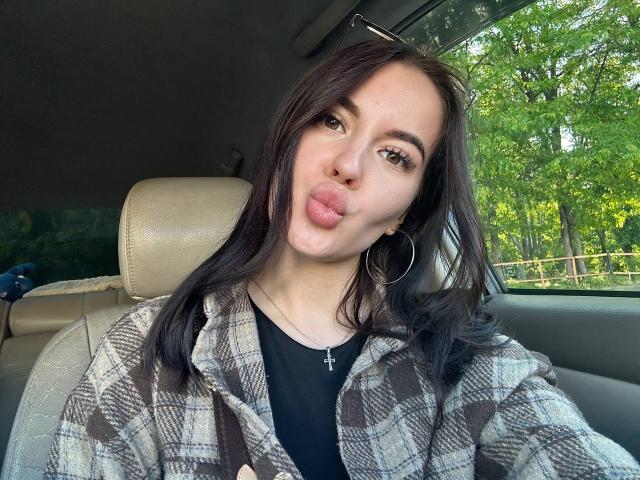 RaichelStoun, puffy lips, black hair, hoop earrings, plaid shirt, black top, cross necklace, car seat, looking at camera, green trees outside, daylight, soft expression.