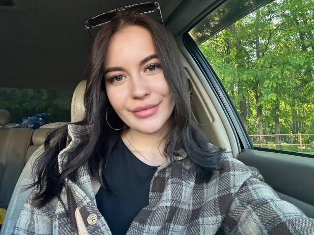 RaichelStoun with dark brown hair, green eyes, and pink lips, wearing a plaid shirt, black top, and hoop earrings, sitting in a car with sunglasses on head.