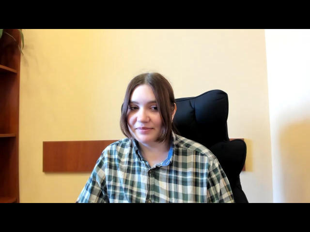 Musiia with shoulder-length brown hair, wearing a green and white plaid shirt, sitting in a black chair, smiling slightly, looking to the right.