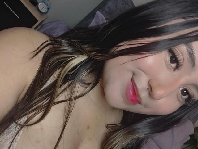 SalomettX, con lunghi capelli neri e biondi, rossetto rosa e un piercing al naso, sorride alla fotocamera, mostrando le spalle scoperte e una piccola imperfezione sul petto..
