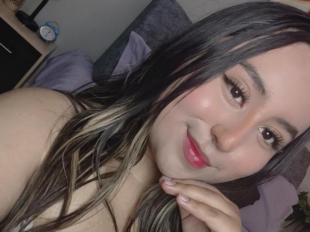 SalomettX has long black hair with blonde highlights, a nose ring, and is smiling with pink lips. She's touching her chin with her right hand. Her tits are not visible.