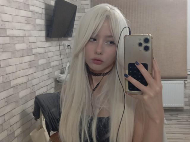 EileneSzala with long white hair, choker, and black dress, holding a phone with earbuds in, taking a mirror selfie, showing blue nails, and a neutral expression.