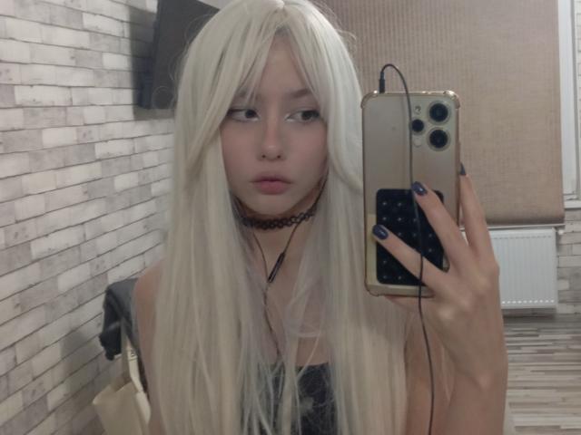 EileneSzala with long blonde hair, pale skin, choker, black nails, holding a gold iPhone, mirror selfie, looking serious, wearing a sheer black top, in a room.