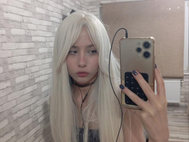 EileneSzala with long white hair, choker, purple nails, holding gold phone, looking at mirror, earbuds in, pale skin, slightly pouty lips, in a room with white brick wall.