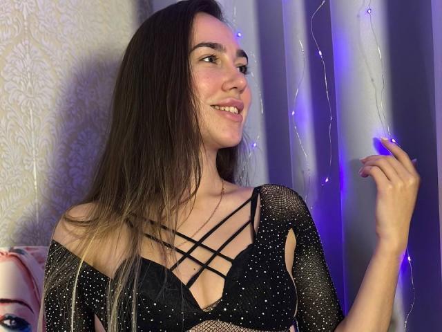 AraiaSheri with long brown hair, smiling, wearing a black mesh top with strappy lingerie, holding a string of purple fairy lights in her right hand.