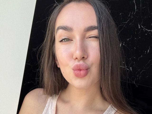 AraiaSheri with long brown hair, winking, pouting her pink lips, green eyes, and wearing a white tank top, against a black marble background.