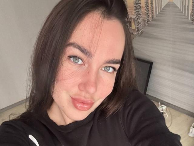 AraiaSheri has pale skin, dark brown hair, blue eyes, and shiny pink lips, wearing a black shirt. She's looking at the camera with a slight smile.