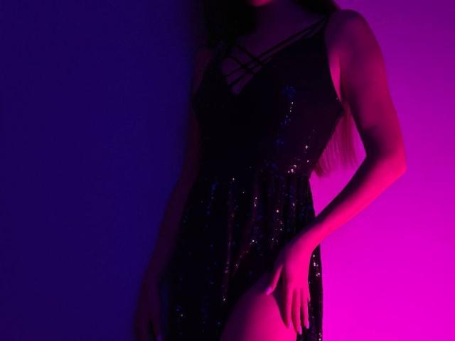 AraiaSheri in a black sequined dress, purple and pink lighting, standing with one hand on her thigh, her ass slightly visible, long brown hair, arm tattoo.