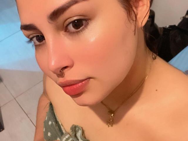 El rostro de SweetSolei69, con piel rosada, ojos marrones, maquillaje sutil y un piercing en la nariz, lleva una blusa verde con un hombro descubierto, mirando hacia arriba con los labios ligeramente entreabiertos y de color rosa..