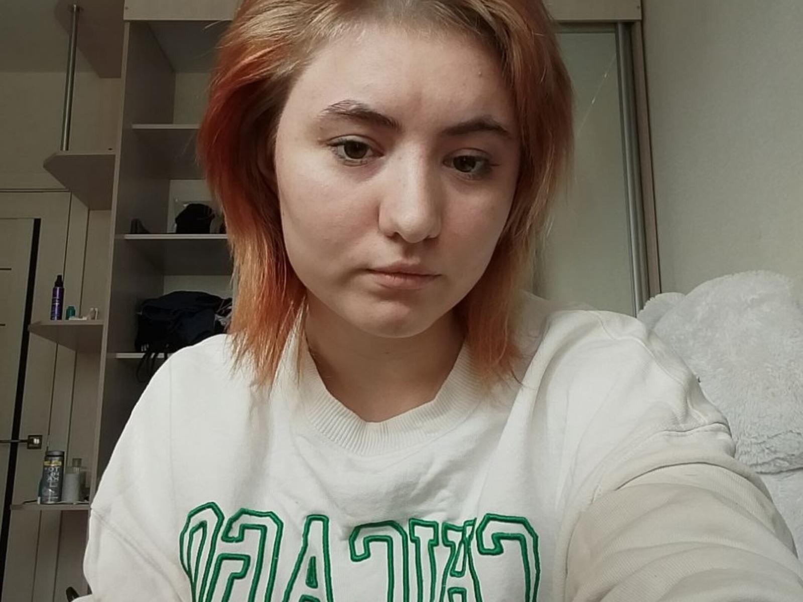 Helene Gilcrease, mit heller Haut, einem kleinen Nasenring und orangefarben gefärbtem Haar, trägt einen grün-weißen Pullover und blickt direkt in die Kamera mit einem neutralen Gesichtsausdruck..