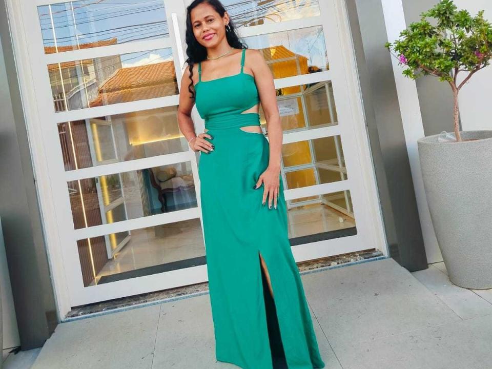 Barbara Lusty indossa un abito verde con tagli, è in piedi, con le mani sui fianchi, mostrando una spaccatura sulla coscia, ha i capelli lunghi e neri, rossetto rosso e un sorriso. Il suo seno è visibile..